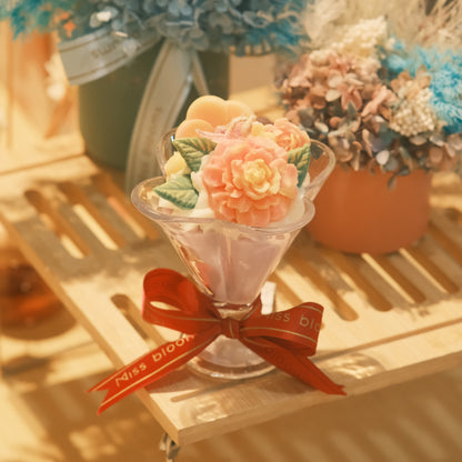 Miss Blooms Flower Cup Candle in martini glass with a large sculptural coral peony wax flower and green leaves, warm golden lighting