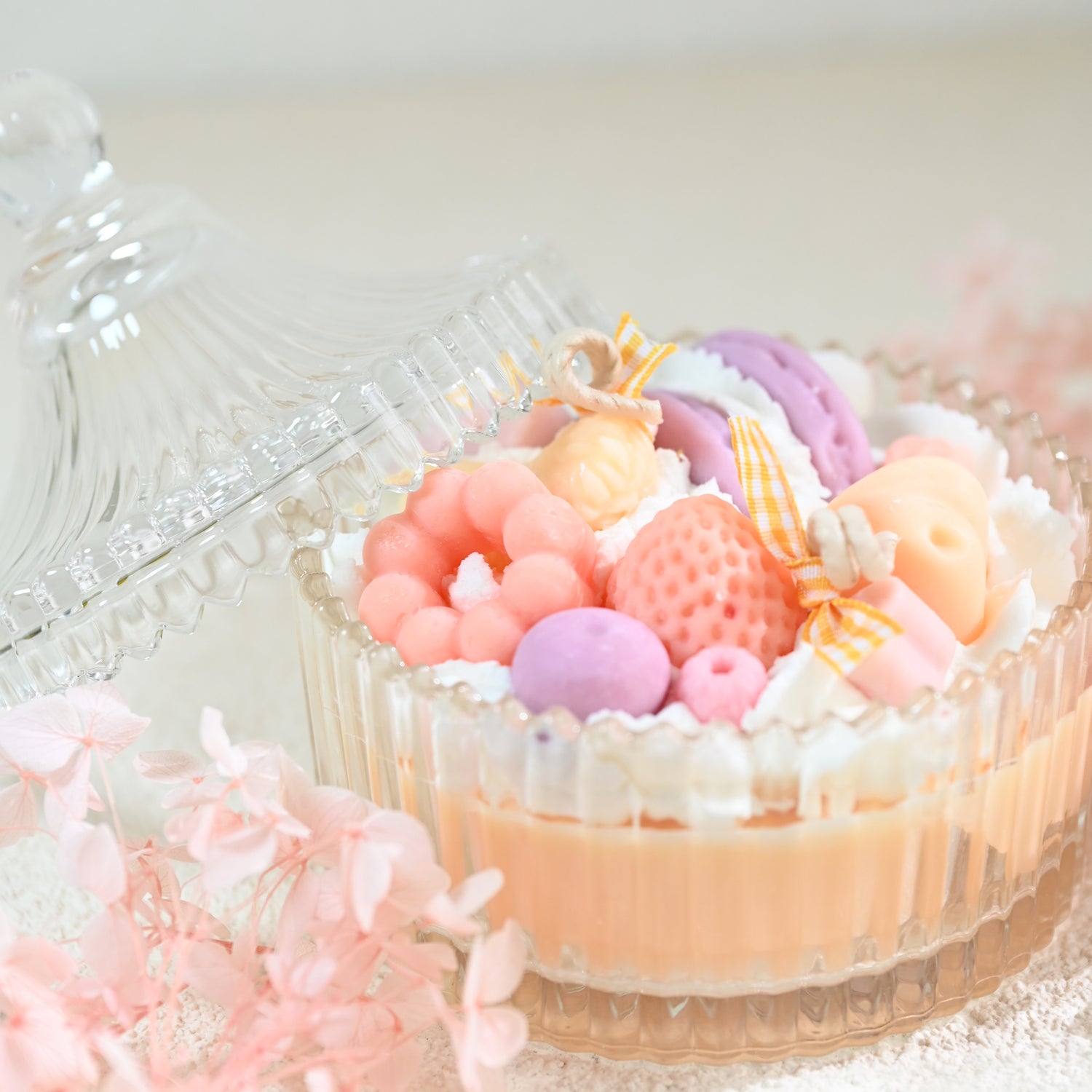 Miss Blooms Sweet Treats Bowl Candle in crystal glass bowl with lid open, showing pastel wax strawberry, macaron and flower decorations with pink flowers in background